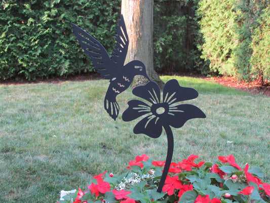 Decorative metal hummingbird and flower sculpture in a garden setting with greenery and flowers.